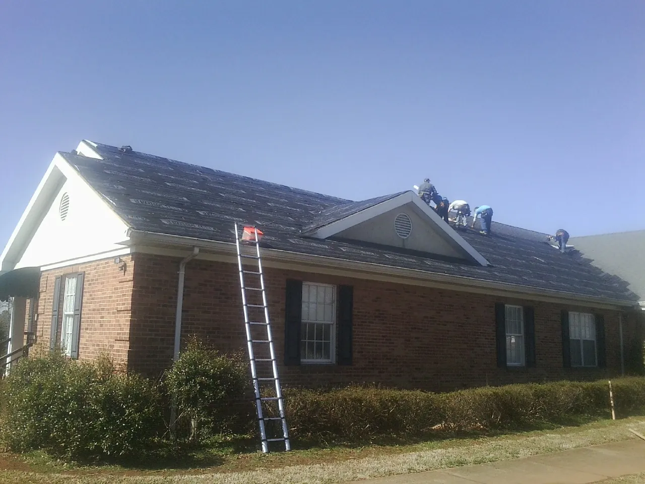 Crew tearing shingles off a brick ranch roof with a ladder set — Emergency Roof Repair work by our crew in Hallwood