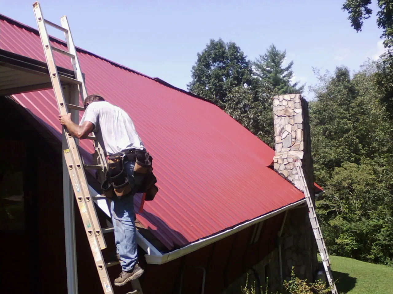 Roofer on a ladder beside a red metal roof and stone chimney — Soffit & Fascia Repair by Roofing Brothers in Hallwood
